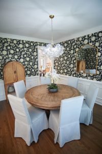 Dining room with dark floral wallpaper, round wood table and slipcovered chairs